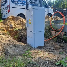 JGinstal usługi elektryczne Jakub Garus - Szara szafka elektryczna INCOBEX na zewnątrz, obok wykopu i pomarańczowego kabla. W tle ogrodzenie, budynek z cegły i auto z napisem 'Usługi Elektryczne'.
