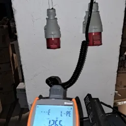 Pomiar napięcia 126.5V na gniazdach siłowych za pomocą miernika MPI-507, widoczne przewody i wtyczki. Ściana z fakturą i kartony w tle.