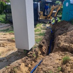 JGinstal usługi elektryczne Jakub Garus - Wkopana niebieska rura w ziemi, obok stoi koparka i słup. Roboty ziemne związane z instalacją podziemną. Widok na zewnątrz, dzień.