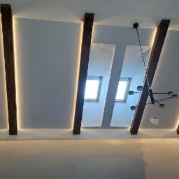 Sufit z drewnianymi belkami i oświetleniem LED, nowoczesny żyrandol. Widoczne okna dachowe i fragment zasłon, sufit po remoncie ze śladami gładzi.