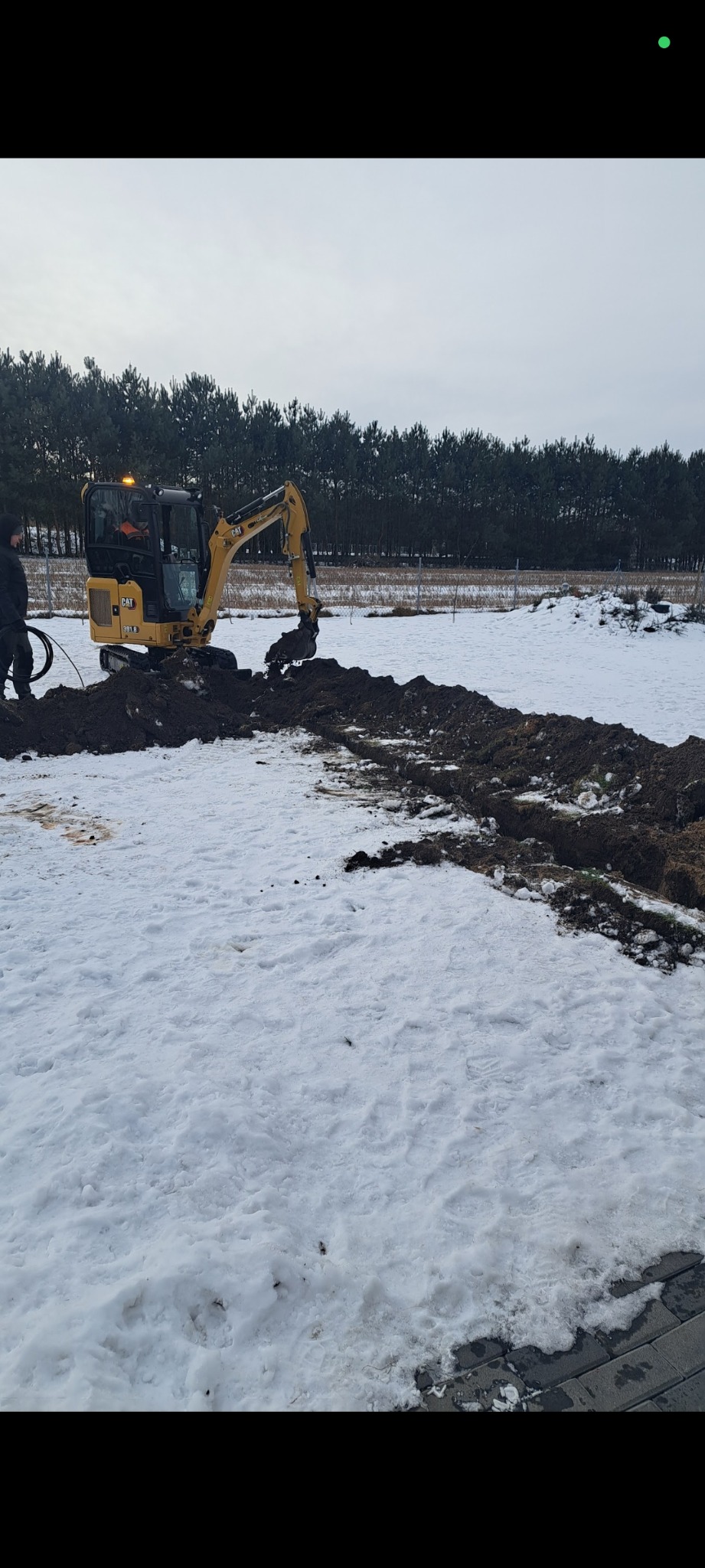 Żółta mini koparka CAT 301.8 kopie rów w zasnieżonym terenie w Pabianicach. Widoczny pracownik i wykopany rów. W tle drzewa.