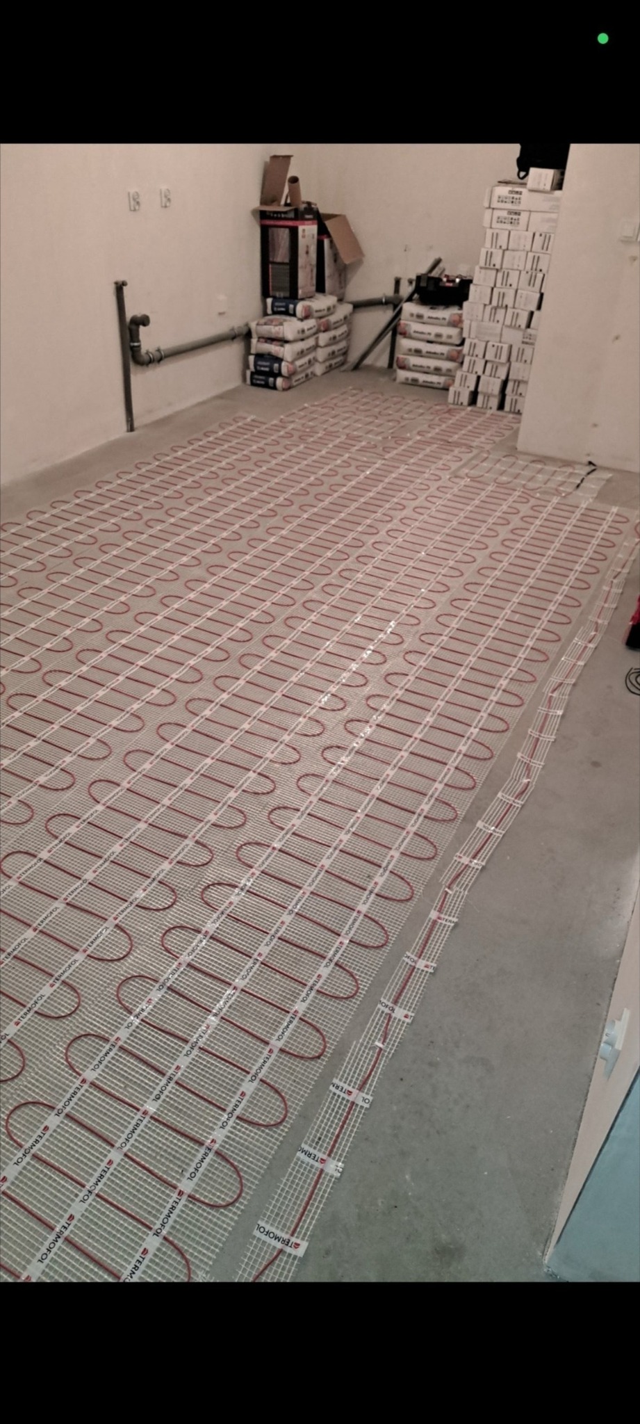 Rozłożona mata grzewcza z czerwonymi przewodami na szarej podłodze w pomieszczeniu, przygotowana do zalania wylewką. W tle worki z zaprawą i kartony.