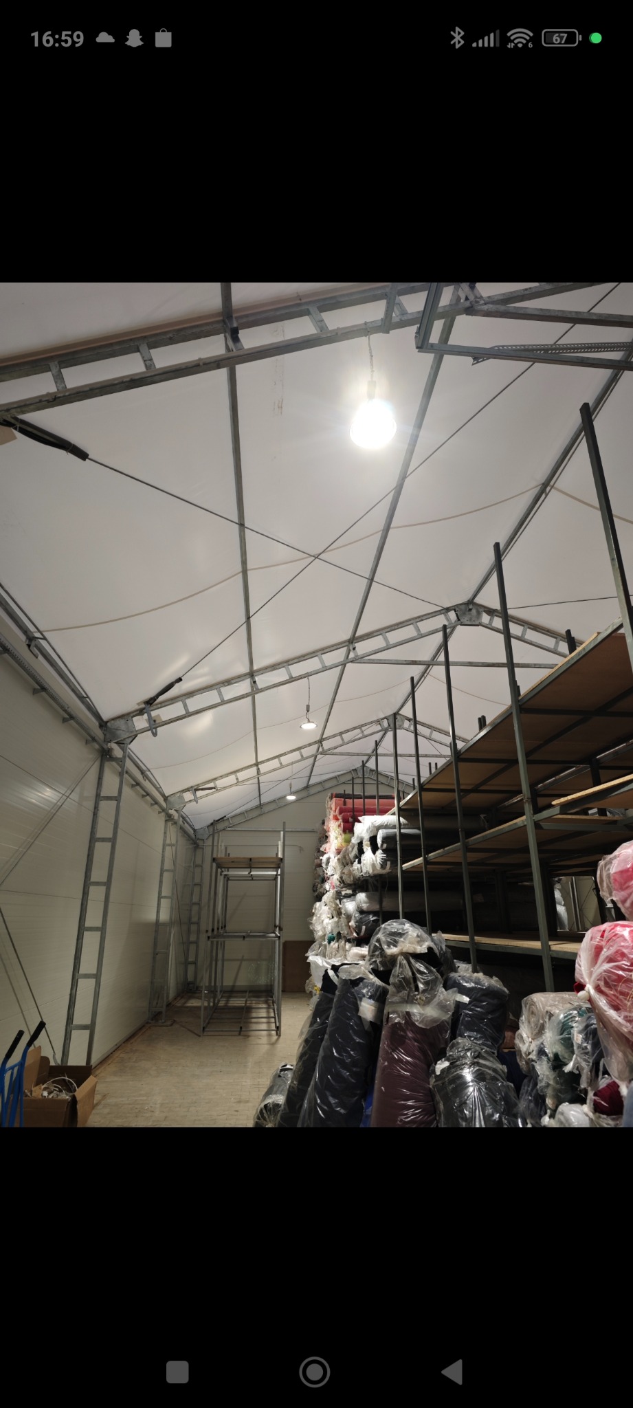 Wnętrze magazynu z metalową konstrukcją dachu, oświetleniem punktowym i regałami wypełnionymi materiałami tekstylnymi w foliowych opakowaniach.