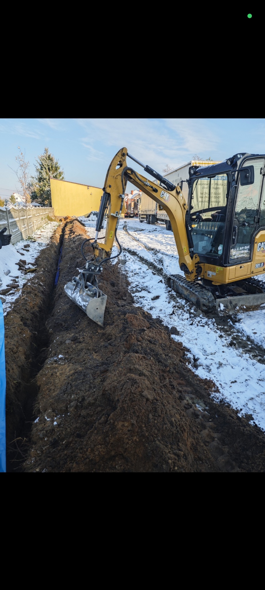 Żółta koparka CAT 301.8 wykopuje rów w ziemi, w którym widać niebieskie rury. Prace ziemne na zewnątrz budynku w zimowej scenerii. Wykop pod przyłącze elektryczne.