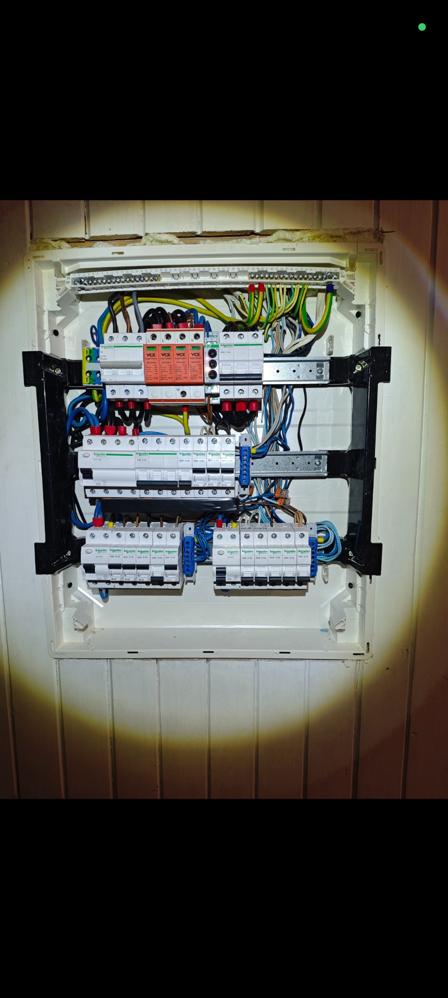 Rozdzielnia elektryczna z zabezpieczeniami przepięciowymi i wyłącznikami różnicowoprądowymi, zamontowana w białej obudowie na ścianie z boazerią.