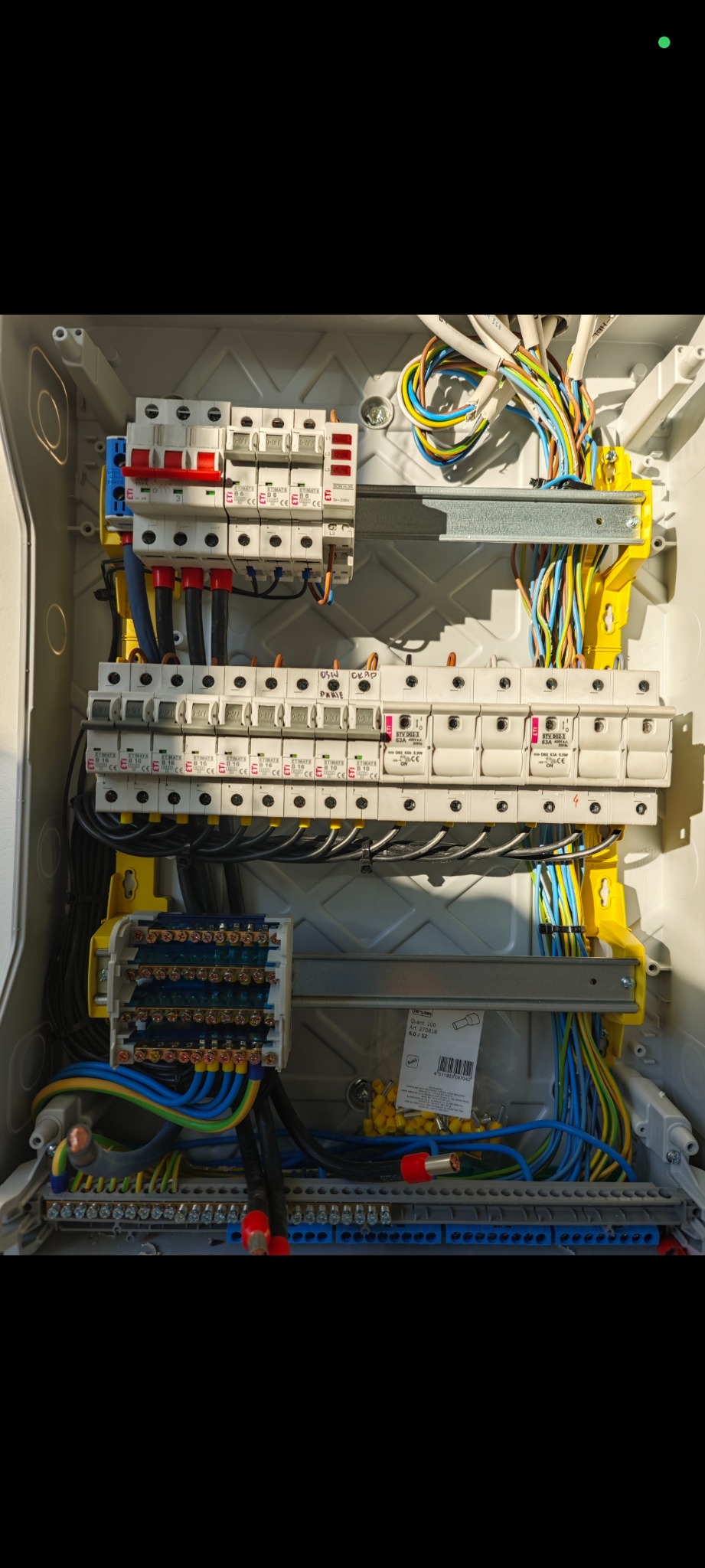 Rozdzielnica elektryczna z bezpiecznikami i przewodami w różnych kolorach, zamontowana w szarej obudowie. Widoczne detale połączeń i oznaczenia.