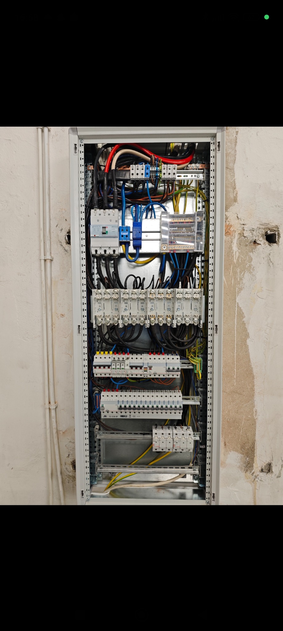 Rozdzielnica elektryczna z licznymi bezpiecznikami, przewodami i elementami montażowymi, umieszczona w szarej obudowie na tle surowej ściany.