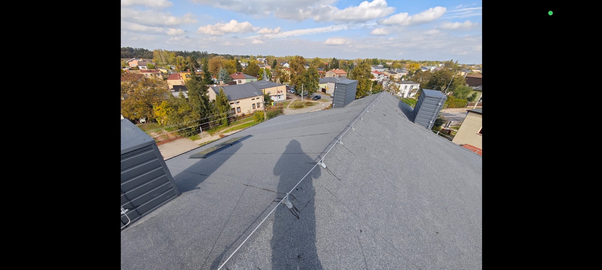 Szary dach z widoczną instalacją odgromową i kominami wentylacyjnymi na tle zabudowy miejskiej w słoneczny dzień. Widoczny cień osoby fotografującej.