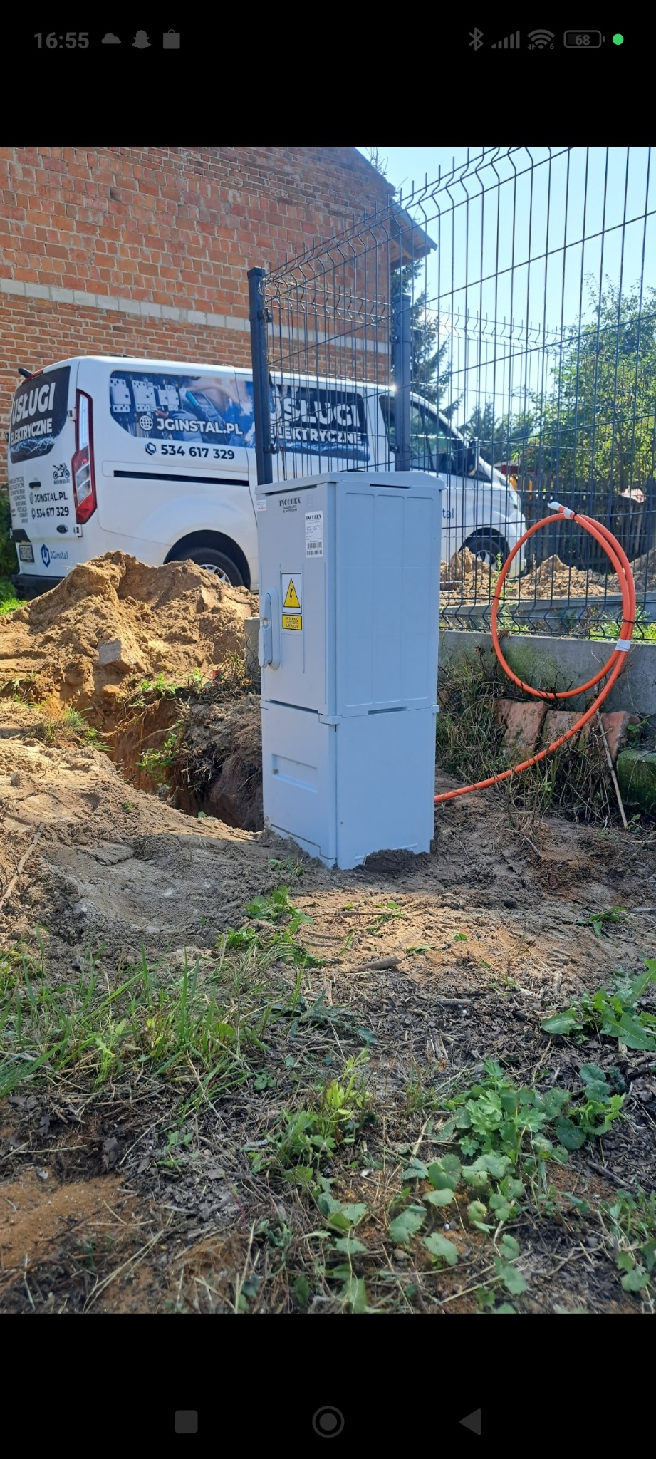 Szara szafka elektryczna INCOBEX na zewnątrz, obok wykopu i pomarańczowego kabla. W tle ogrodzenie, budynek z cegły i auto z napisem 'Usługi Elektryczne'.