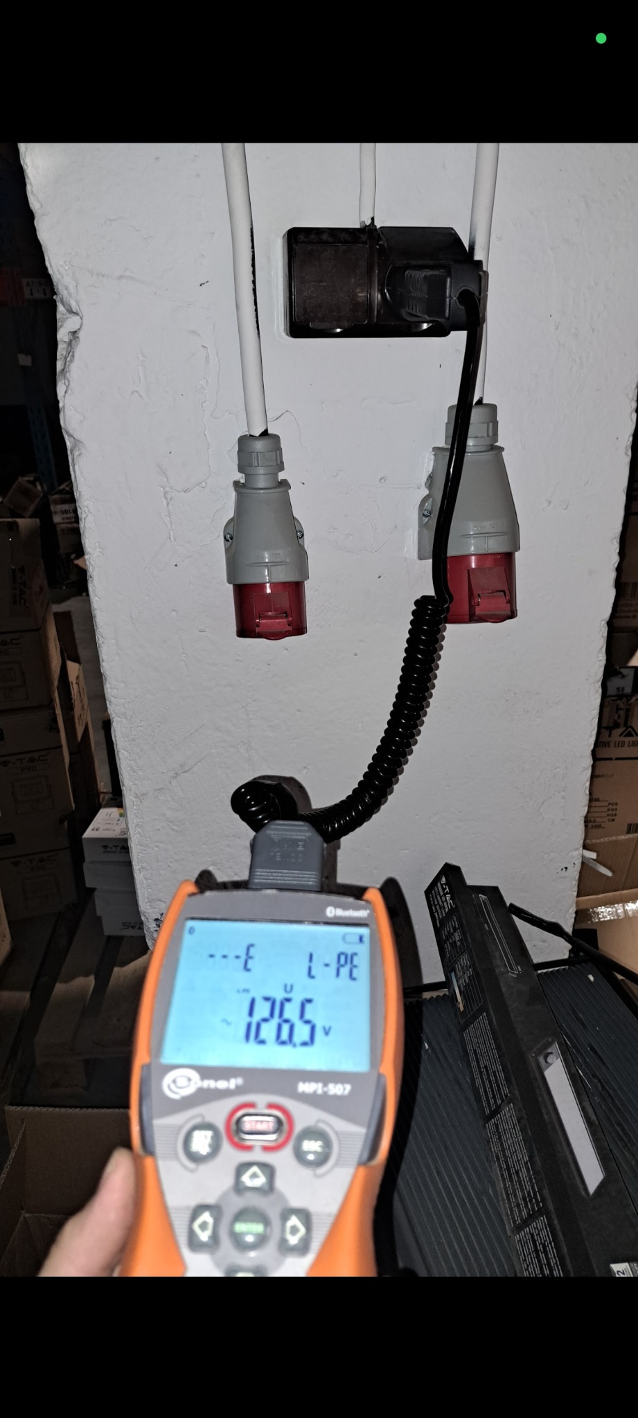 Pomiar napięcia 126.5V na gniazdach siłowych za pomocą miernika MPI-507, widoczne przewody i wtyczki. Ściana z fakturą i kartony w tle.