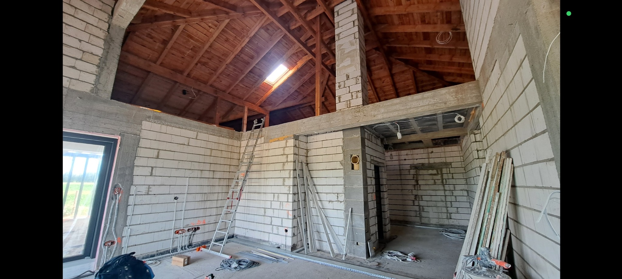 Wnętrze domu w trakcie budowy z widoczną drewnianą konstrukcją dachu, ścianami z białego bloczka, instalacją elektryczną i rozłożonymi narzędziami.