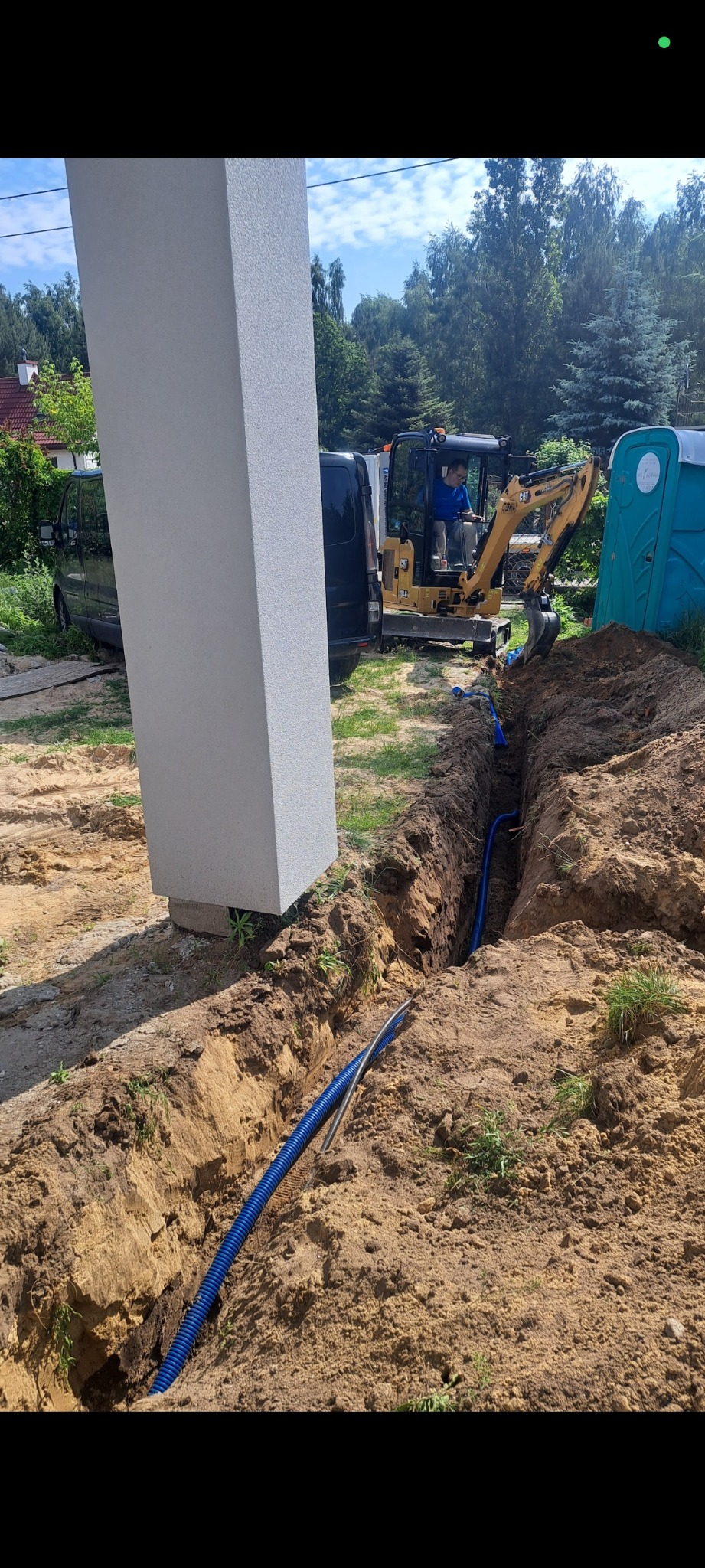 Wkopana niebieska rura w ziemi, obok stoi koparka i słup. Roboty ziemne związane z instalacją podziemną. Widok na zewnątrz, dzień.