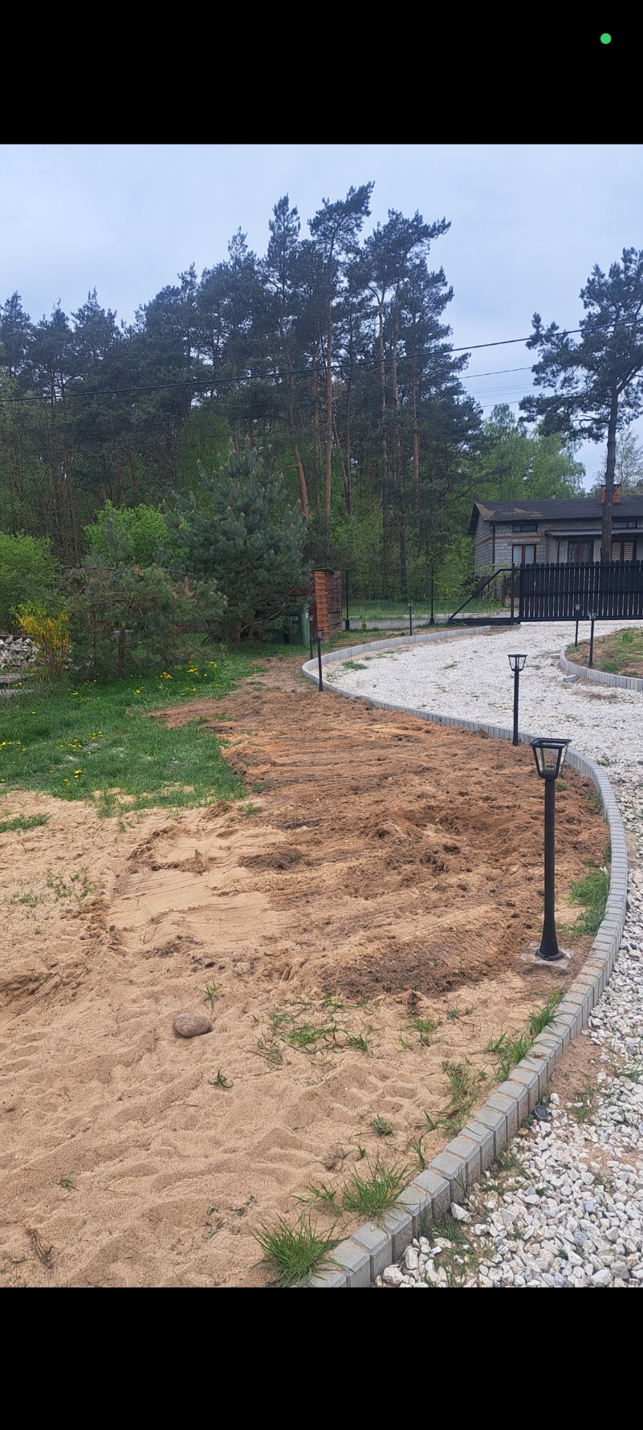 Ogród w trakcie prac ziemnych, widoczne lampy ogrodowe wzdłuż wytyczonej ścieżki, w tle drzewa i budynek. Przygotowanie terenu pod trawnik/nasadzenia.