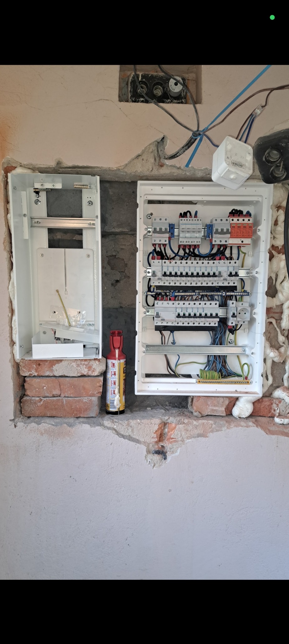 Otwarta, biała skrzynka rozdzielcza z zainstalowanymi bezpiecznikami i przewodami, zamontowana w surowej ścianie z widoczną cegłą i pianką montażową.