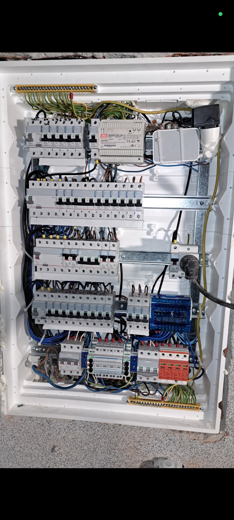 Rozdzielnia elektryczna z licznymi zabezpieczeniami, okablowaniem, modułami i listwami, umieszczona w białej obudowie. Widok z góry na instalację wewnątrz.