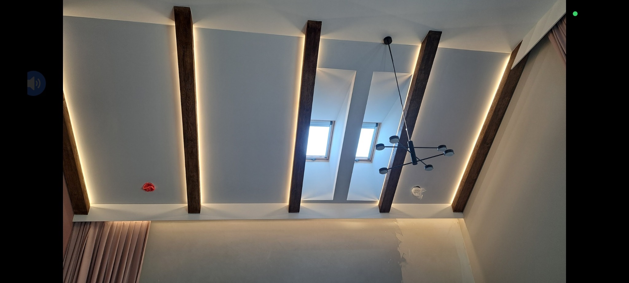 Sufit z drewnianymi belkami i oświetleniem LED, nowoczesny żyrandol. Widoczne okna dachowe i fragment zasłon, sufit po remoncie ze śladami gładzi.