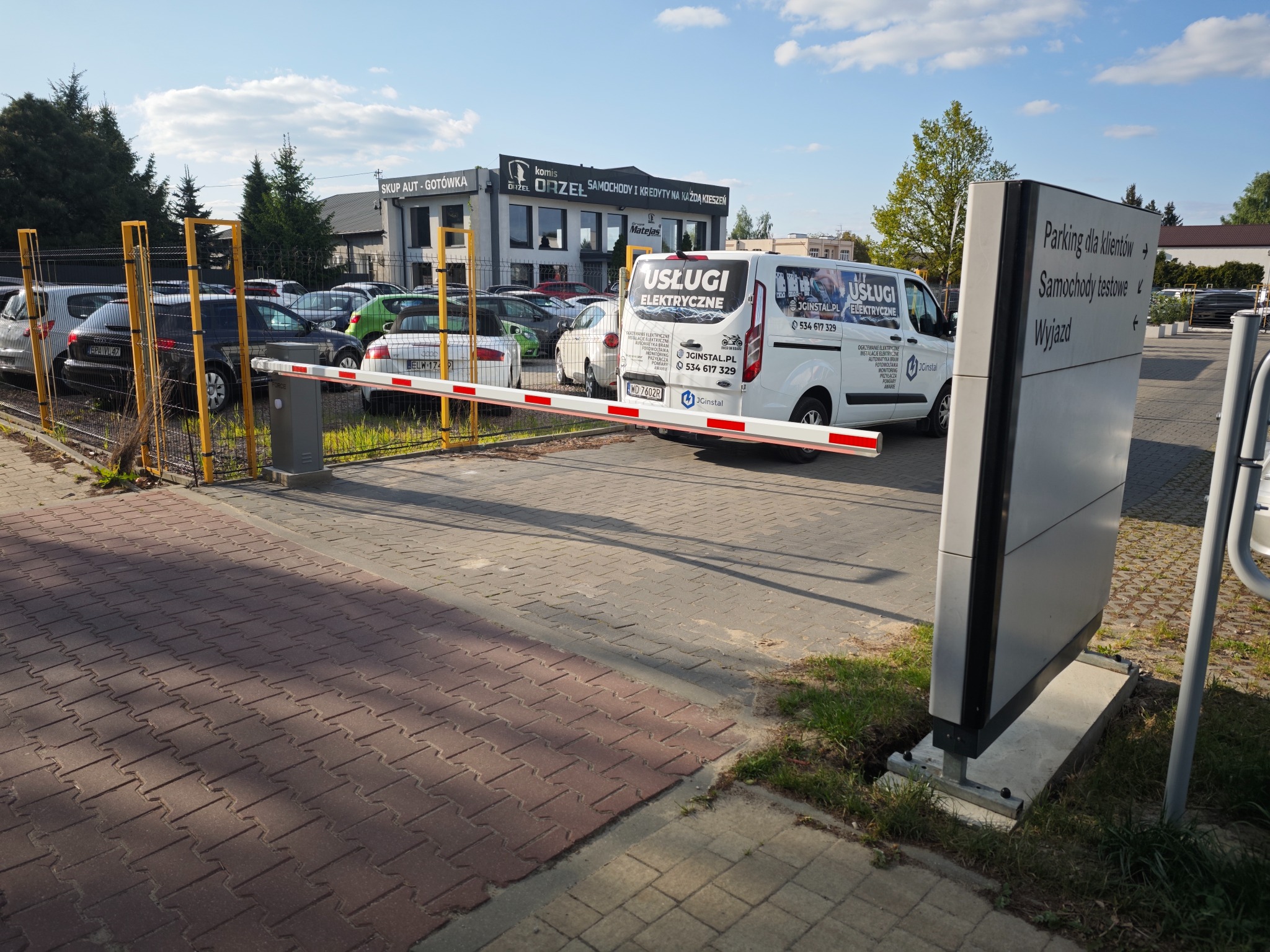 Van z reklamą usług elektrycznych parkuje na parkingu. W tle budynek komisu samochodowego i szlaban. Tablica informacyjna wskazuje kierunek do parkingu.