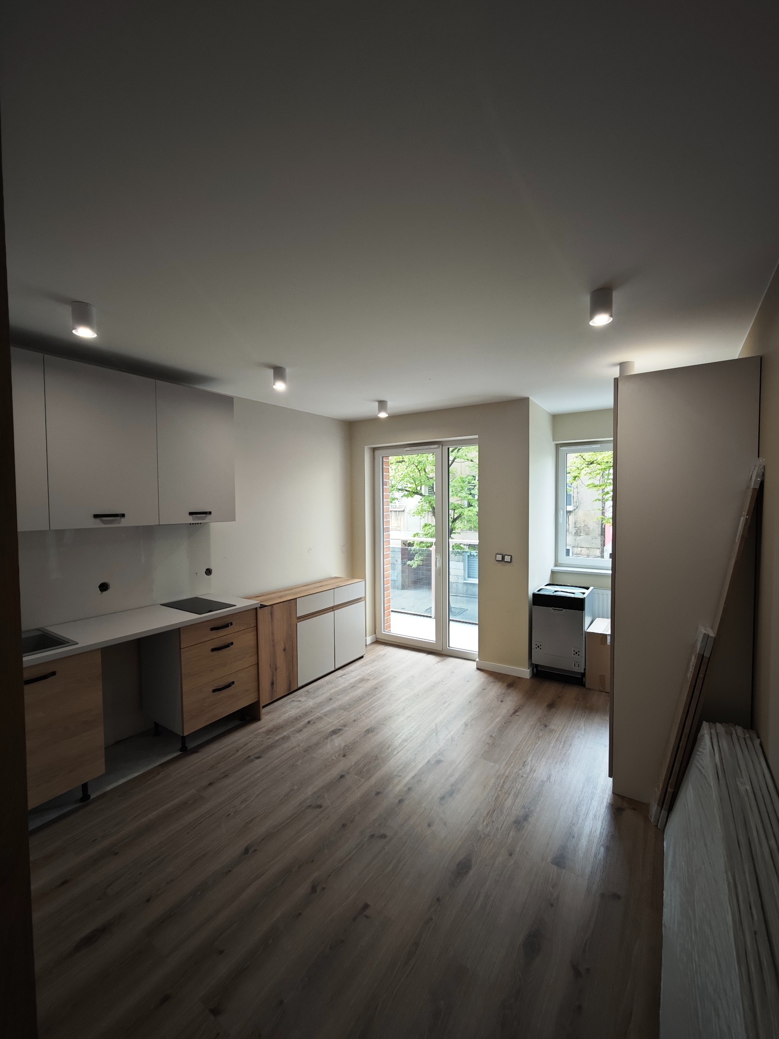 Jasne wnętrze mieszkania po remoncie: drewniana podłoga, biało-drewniana zabudowa kuchenna, okna balkonowe z widokiem na zieleń i punktowe oświetlenie sufitowe.