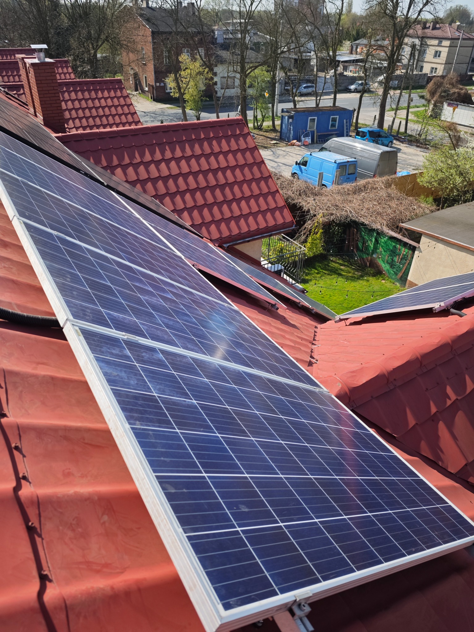 Panele słoneczne na czerwonym dachu domu, widok z góry, z elementami zabudowy w tle, słoneczny dzień. Detal montażu paneli.