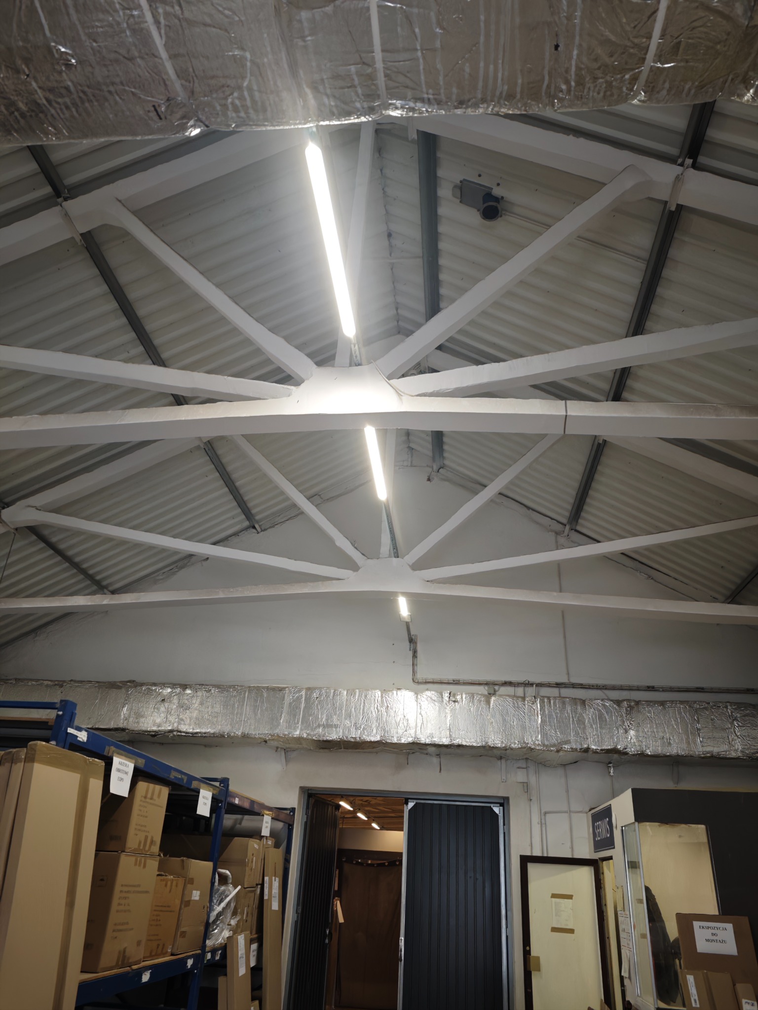 Biały sufit magazynu z widocznymi lampami liniowymi, metalową konstrukcją i fragmentem instalacji pod sufitem. Widoczny regał z kartonami i brama.
