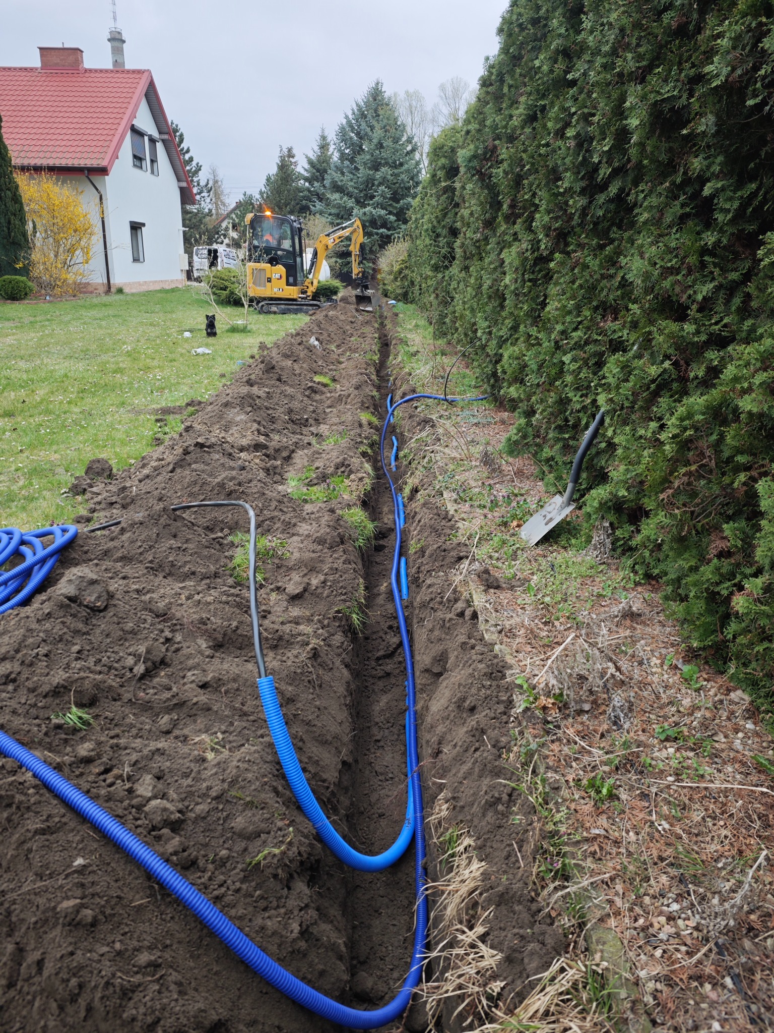 Wykop z ułożonymi niebieskimi rurami karbowanymi, prawdopodobnie peszel, w tle koparka i budynek mieszkalny. Prace ziemne i instalacyjne w toku.