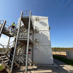 WPTechnik Karol Fąfara - Biały, modułowy budynek z trzema klimatyzatorami na ścianie, metalowe schody z pracownikami wchodzącymi po nich. Błękitne niebo w tle.