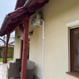 Biała pompa ciepła na elewacji domu z brązową stolarką okienną i zadaszeniem tarasu. Widoczne elementy montażowe i rynna odprowadzająca wodę. Dzień, pochmurno.