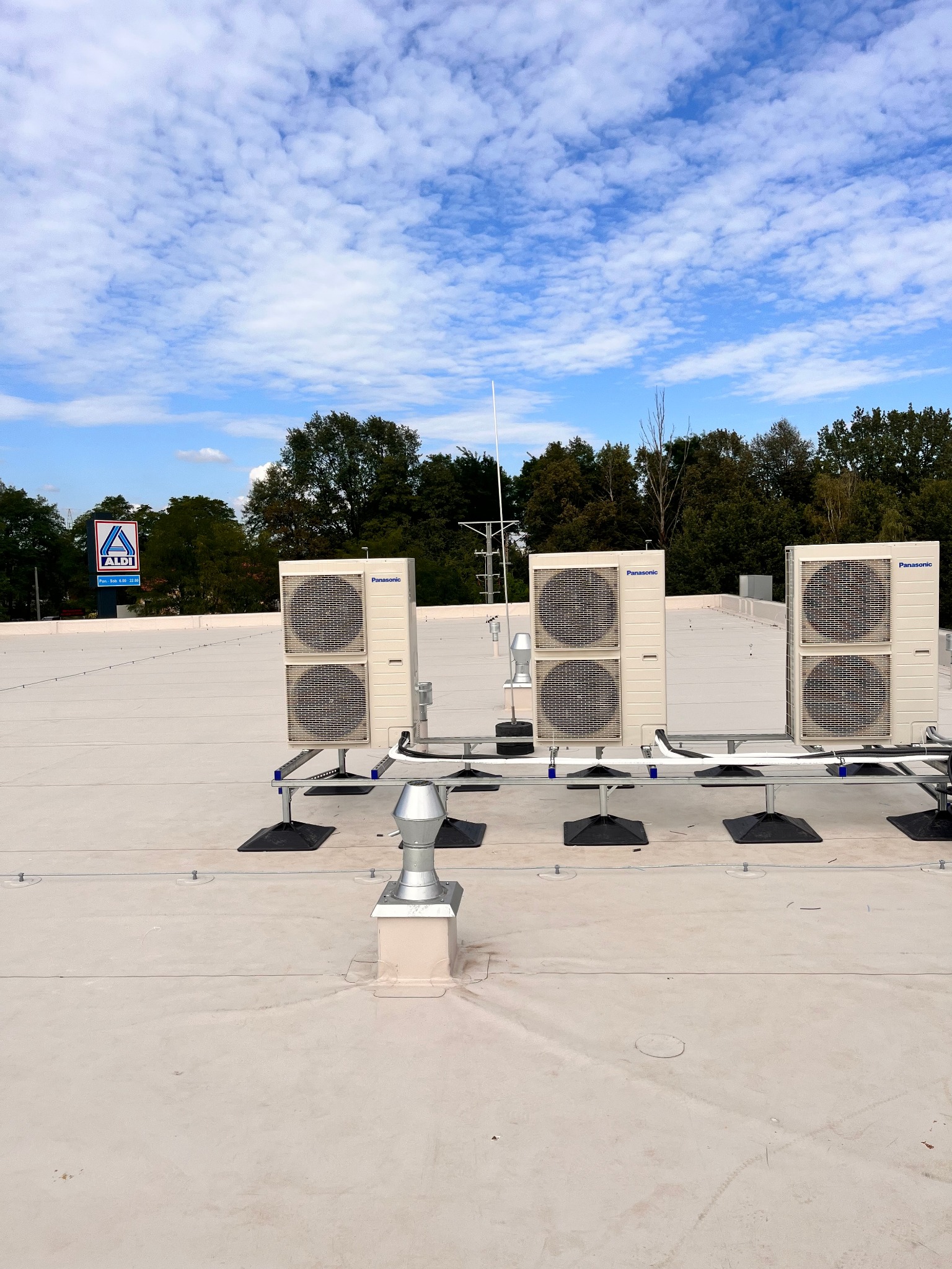 Trzy jednostki zewnętrzne klimatyzacji Panasonic zamontowane na dachu budynku z widocznym znakiem Aldi w tle, instalacja na metalowych konstrukcjach wsporczych.