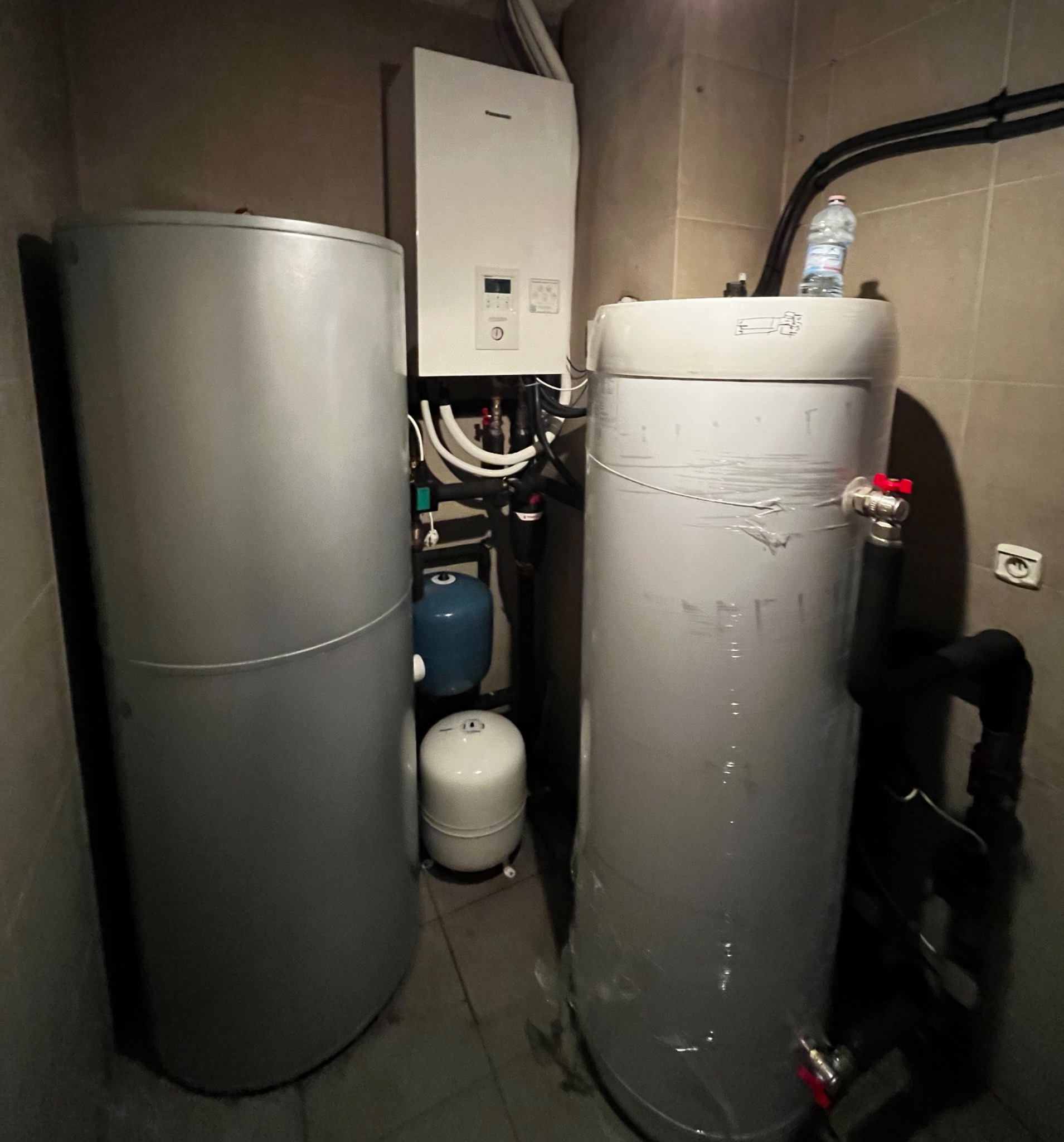 Instalacja w kotłowni z dwoma srebrnymi zbiornikami, białym modułem sterującym na ścianie i niebieskim zbiornikiem wyrównawczym, z butelką wody na górze jednego ze zbiorników.