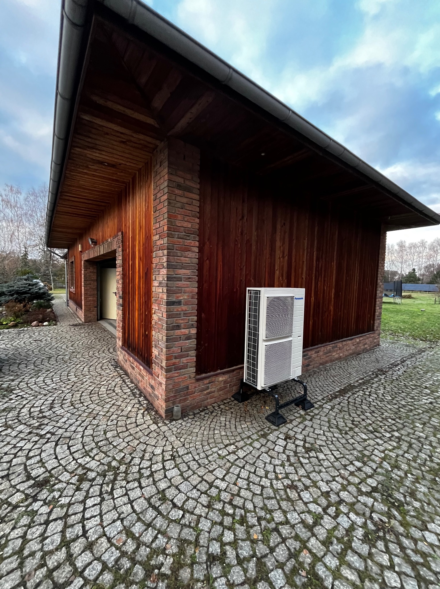 Zewnętrzna jednostka klimatyzacyjna Panasonic ustawiona na czarnym stelażu przed budynkiem z drewnianą elewacją i ceglanymi narożnikami, na brukowanym podłożu.