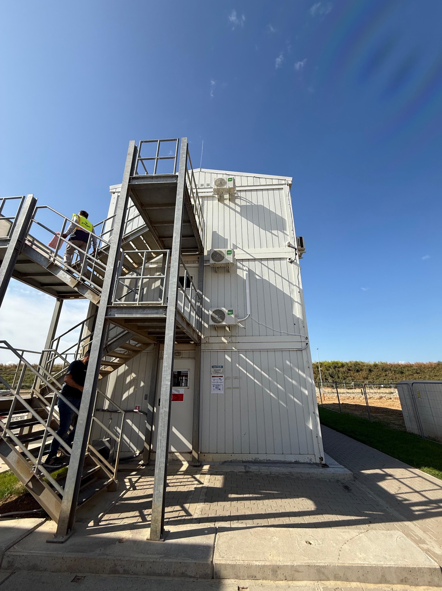Biały, modułowy budynek z trzema klimatyzatorami na ścianie, metalowe schody z pracownikami wchodzącymi po nich. Błękitne niebo w tle.
