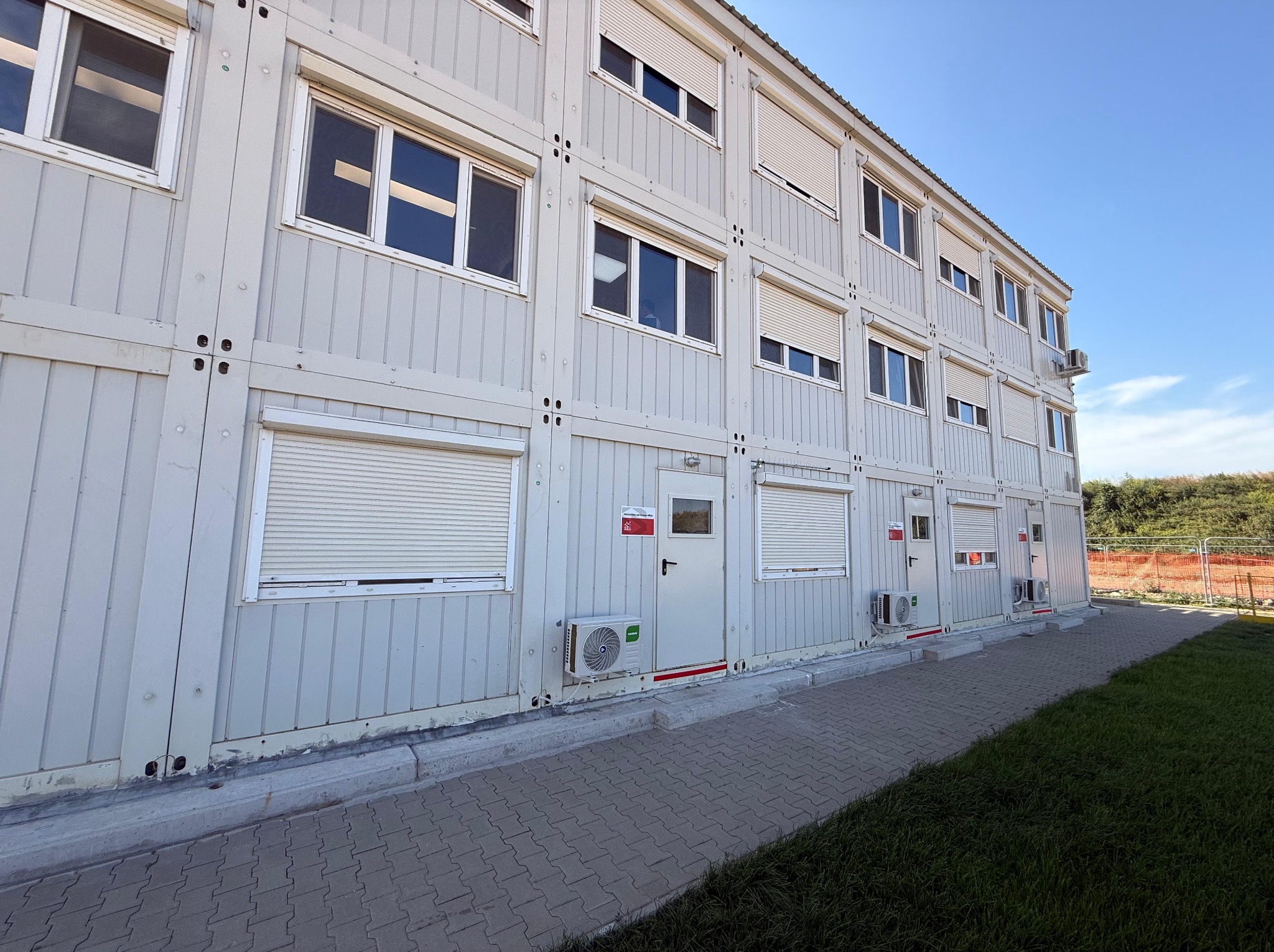 Dwukondygnacyjny budynek modułowy z klimatyzacją i roletami zewnętrznymi, widziany z dołu pod kątem. Szara kostka brukowa i trawnik na pierwszym planie.