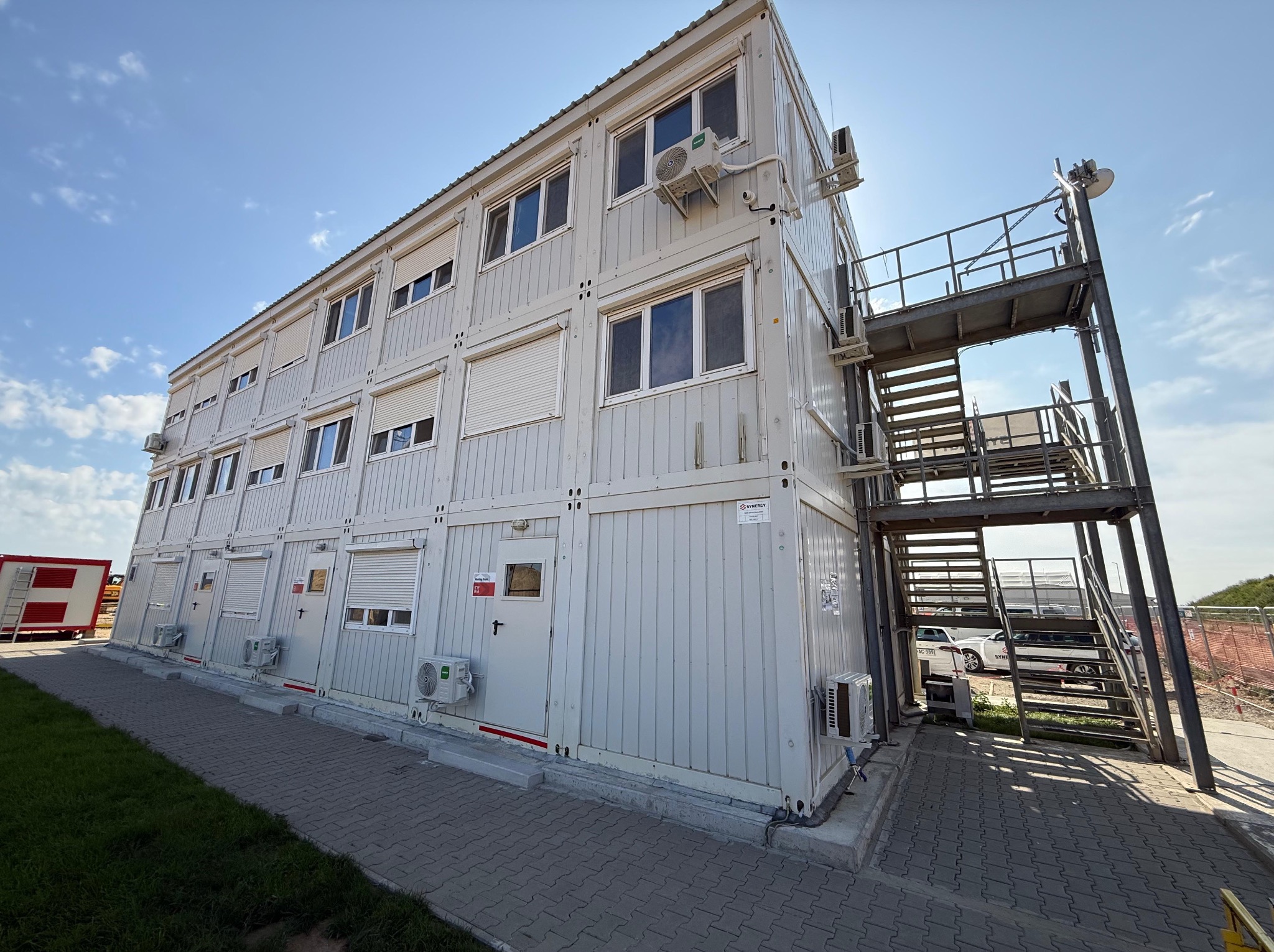 Dwukondygnacyjny budynek modułowy z klimatyzacją, schodami zewnętrznymi i widocznymi detalami konstrukcyjnymi na tle błękitnego nieba.