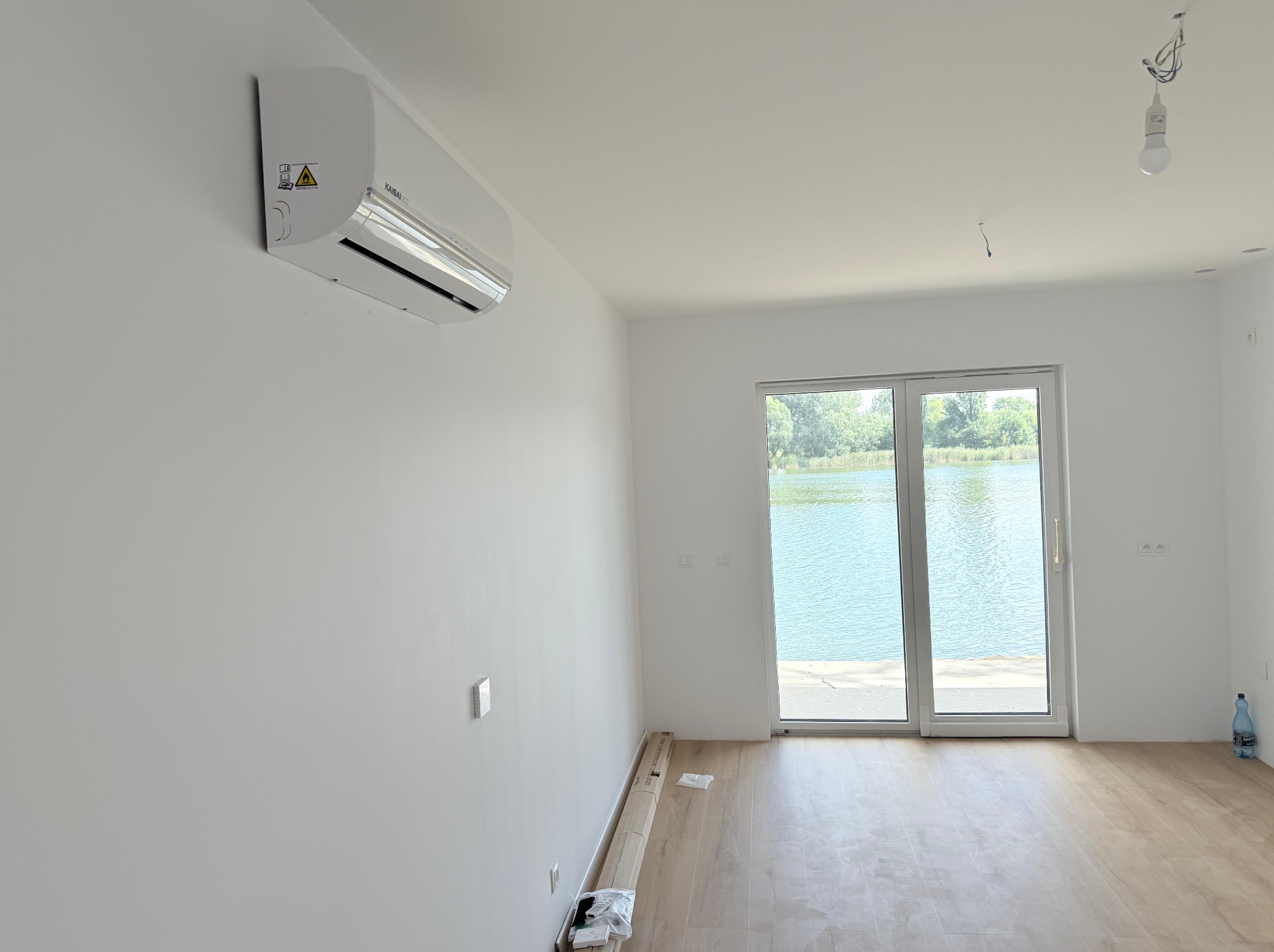 Biała klimatyzacja Kaisai Eco zamontowana w jasnym, minimalistycznym wnętrzu z widokiem na jezioro przez przesuwne drzwi balkonowe. Podłoga w panelach, surowa żarówka.