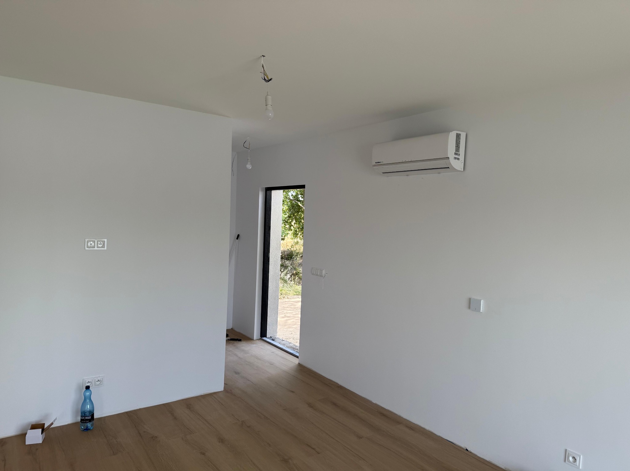 Wnętrze z zamontowaną klimatyzacją na białej ścianie, drewniana podłoga, widoczne gniazdka i instalacja elektryczna, surowy stan wykończenia.