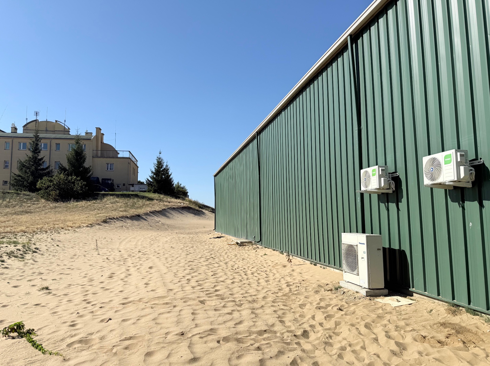 Instalacja klimatyzacji: trzy jednostki zewnętrzne na zielonej, blaszanej ścianie budynku, piasek na pierwszym planie, budynek w tle, słoneczny dzień.