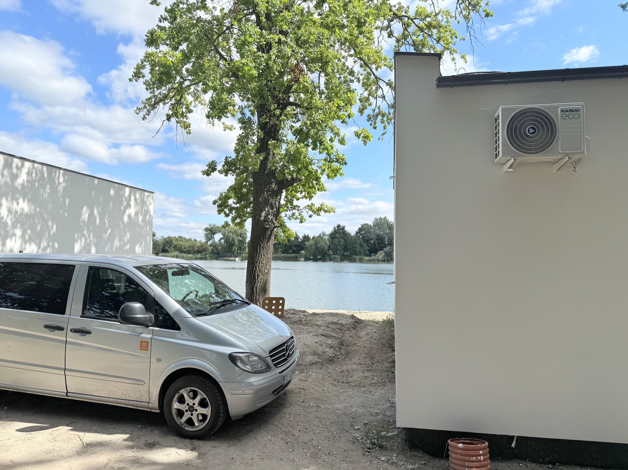 Biała pompa ciepła Kaisai Eco zamontowana na ścianie budynku, obok zaparkowany srebrny van z widokiem na jezioro i drzewa w słoneczny dzień.