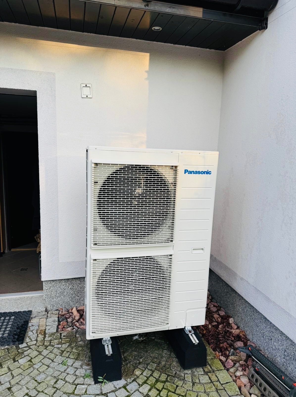 Biała, dwuczęściowa pompa ciepła Panasonic na czarnych podstawach, zainstalowana przy jasnej ścianie budynku z kostką brukową i kamieniami ozdobnymi.