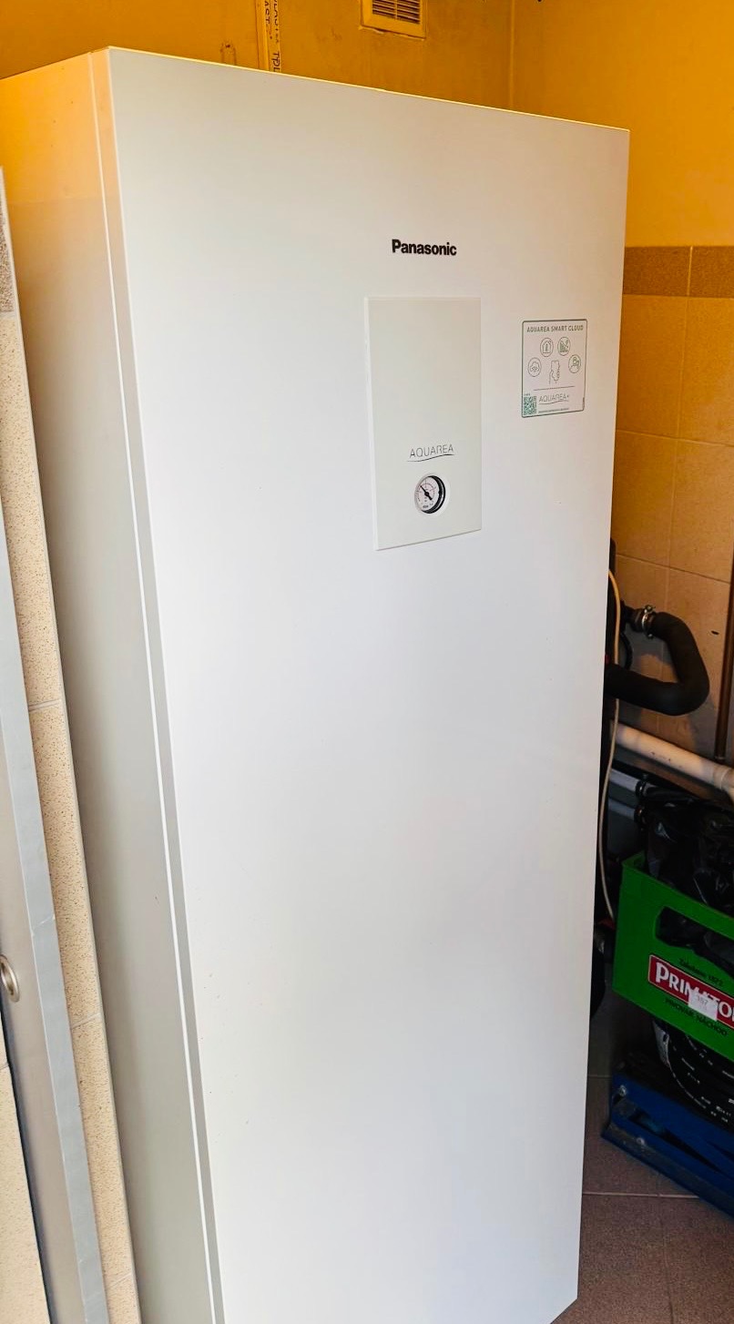 Biała pompa ciepła Panasonic Aquarea w pomieszczeniu z beżowymi płytkami, widoczne przyłącza i skrzynka. Urządzenie sprawne i gotowe do użycia.