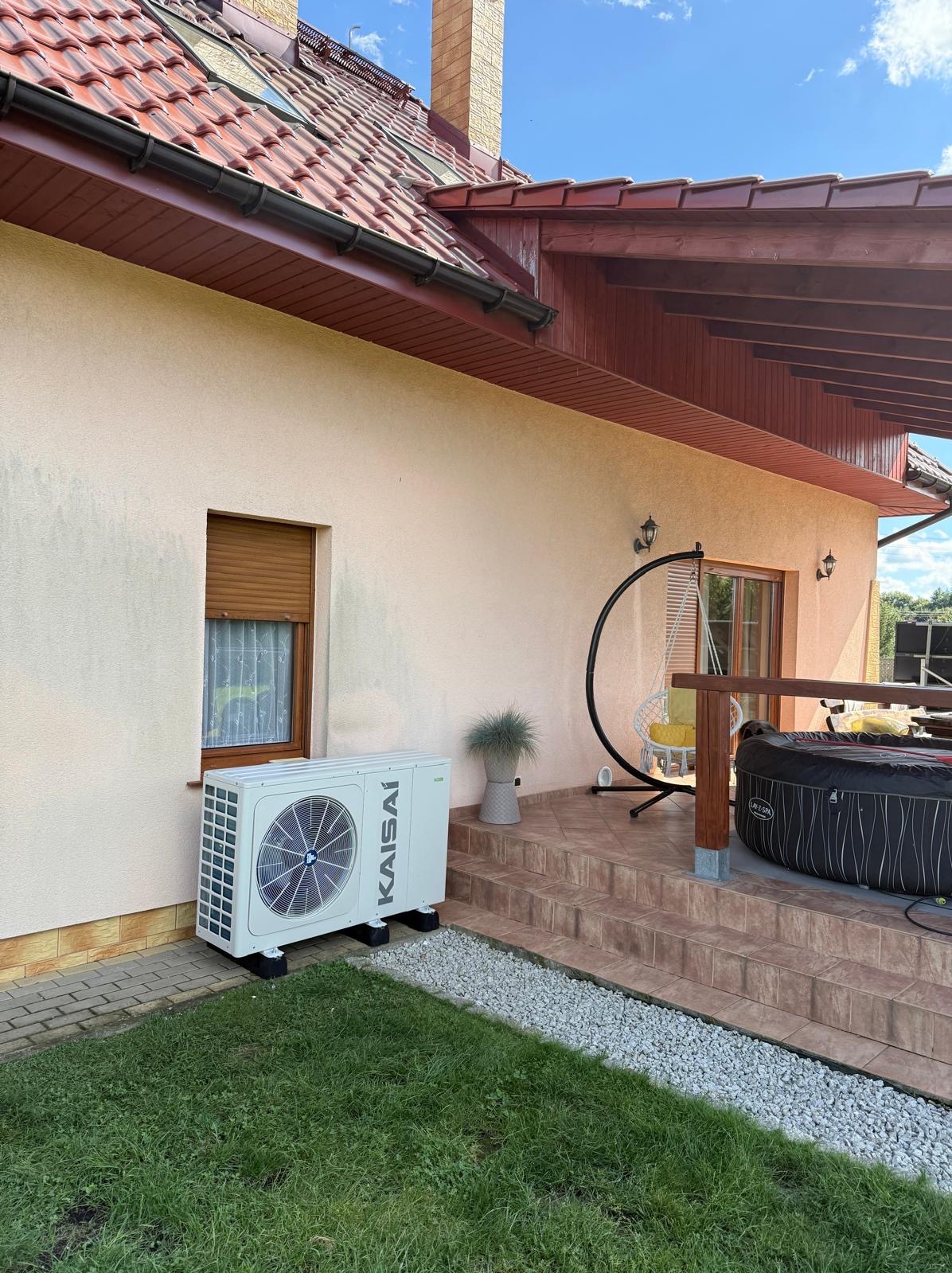 Biała pompa ciepła Kaisai zamontowana przy beżowej ścianie domu z brązową stolarką okienną i tarasem z jacuzzi. Widok na trawnik i błękitne niebo.