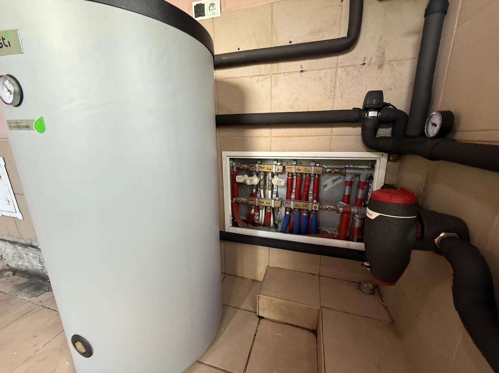 Biały zasobnik ciepłej wody użytkowej, obok rozdzielacz centralnego ogrzewania z rurami w izolacji. Instalacja w kotłowni z płytkami na ścianach.