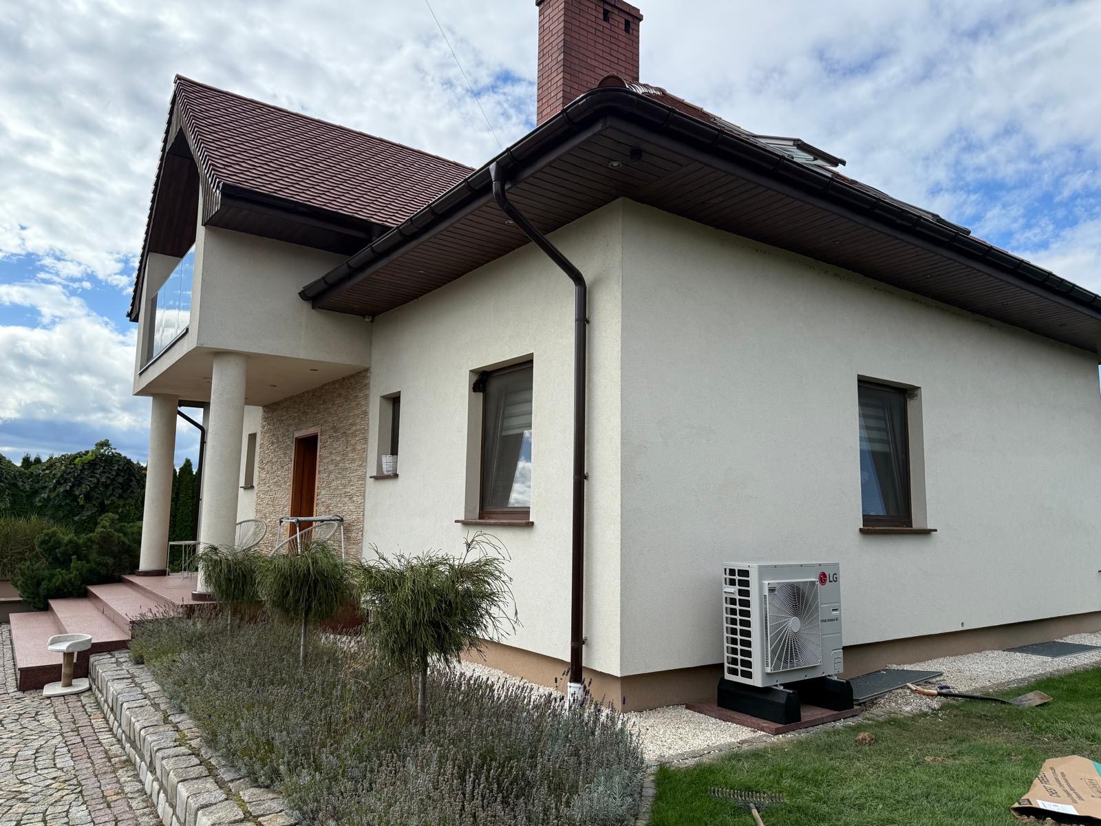 Biały dom jednorodzinny z zainstalowaną pompą ciepła LG Therma V na zewnątrz, widok z boku, zieleń w tle.