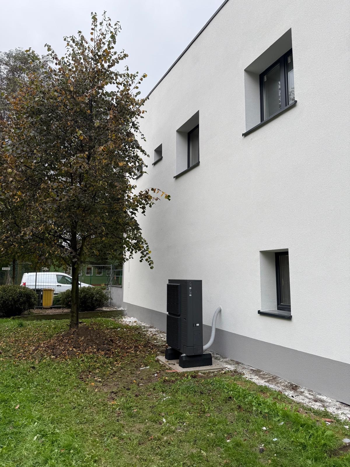Szara pompa ciepła zamontowana przy białej ścianie budynku z małymi oknami, obok drzewo z brązowymi liśćmi, trawnik i fragment zaparkowanego samochodu.