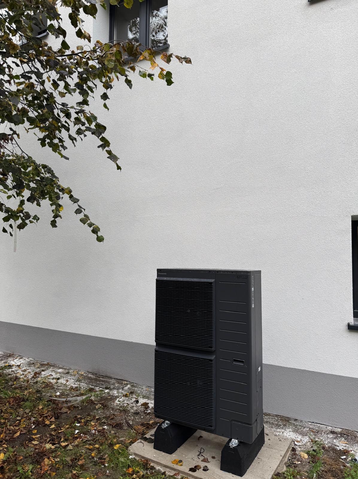 Czarna pompa ciepła Panasonic Aquarea, umieszczona na betonowej podstawie przy białej ścianie budynku. Widoczne liście na ziemi i gałąź drzewa w lewym górnym rogu.
