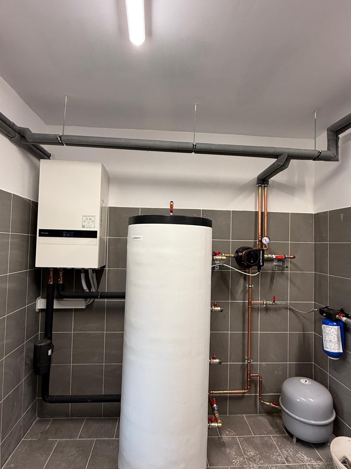 Zainstalowana biała pompa ciepła z zasobnikiem w kotłowni z szarymi płytkami. Widoczne rury miedziane, czarne izolowane oraz filtr wody. Oświetlenie górne.