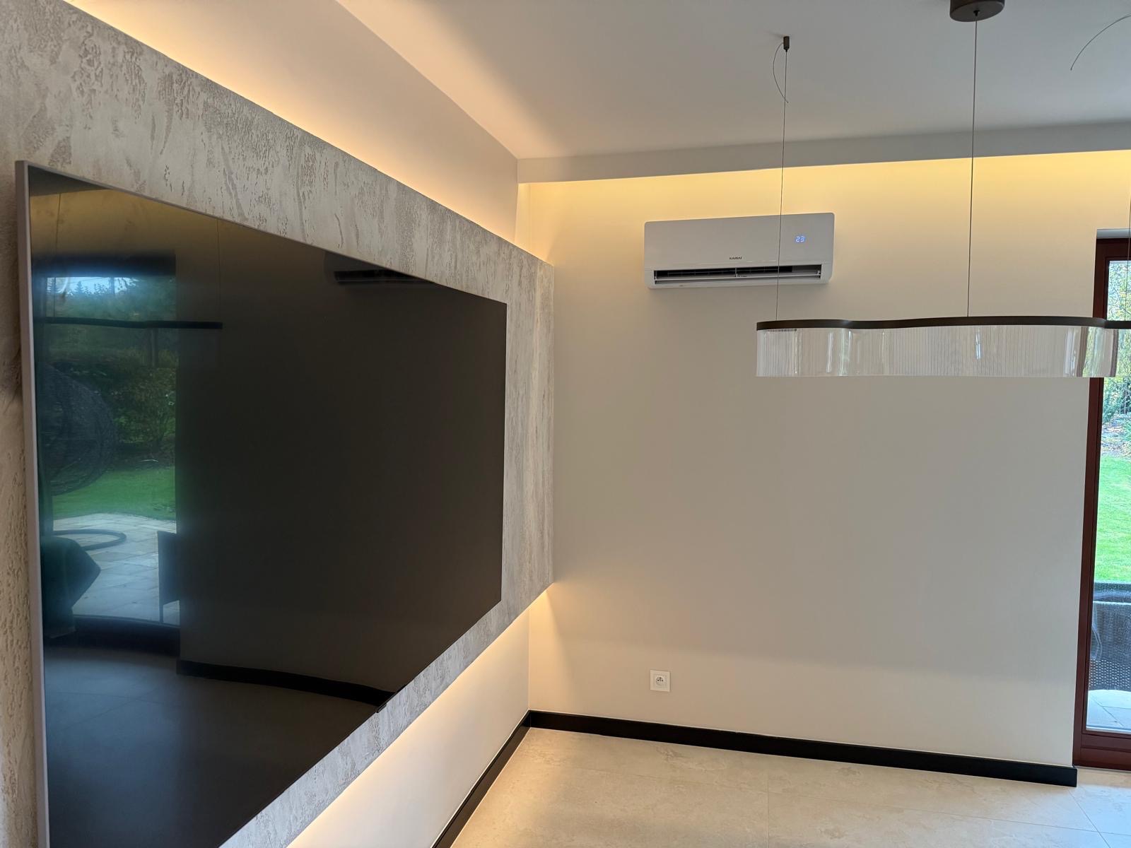 Nowoczesne wnętrze z klimatyzatorem ściennym, podświetlaną ścianą dekoracyjną i minimalistyczną lampą wiszącą. Widoczny fragment okna i ogrodu.