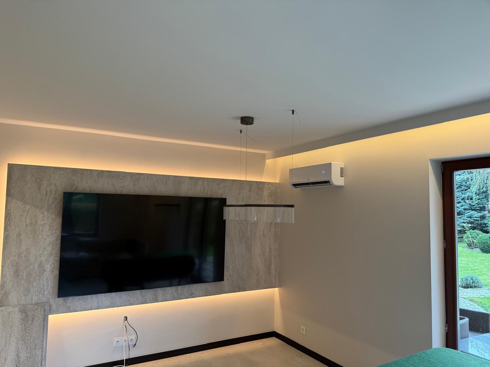 Nowoczesne wnętrze z klimatyzatorem ściennym, podświetleniem LED i telewizorem na tle ściany z teksturą. Widok na zewnątrz przez okno.