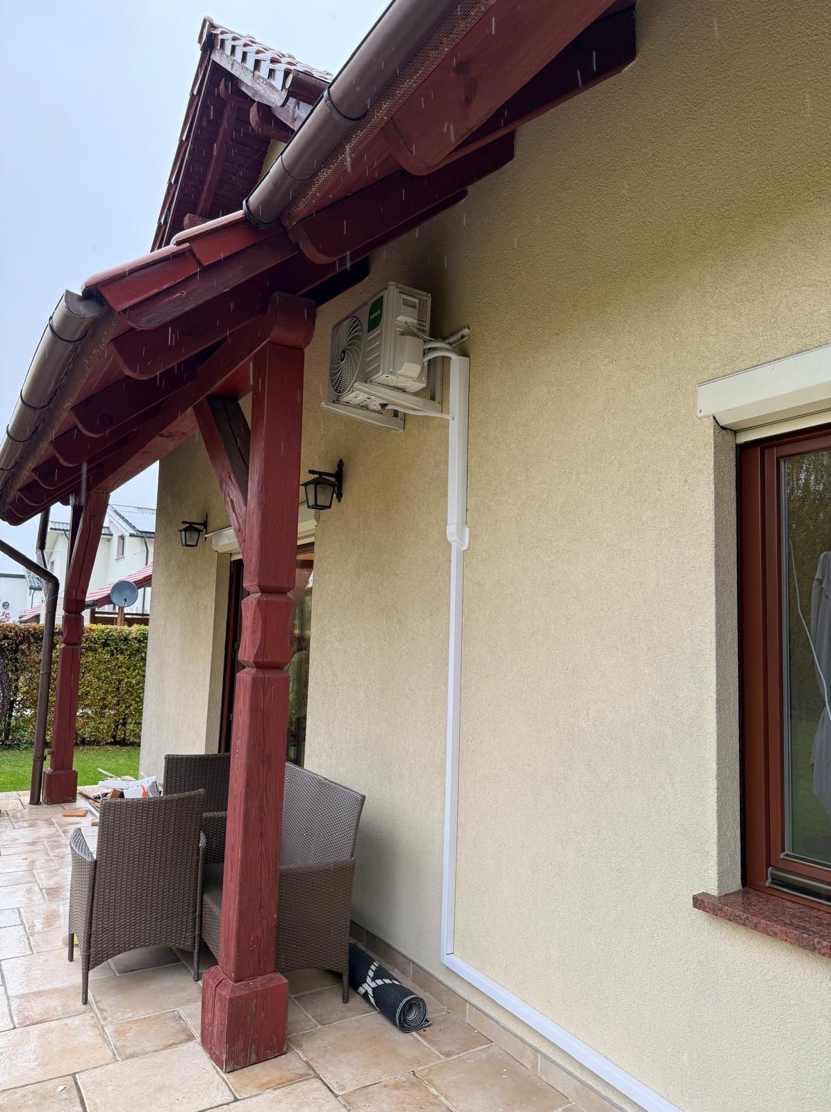 Biała pompa ciepła na elewacji domu z brązową stolarką okienną i zadaszeniem tarasu. Widoczne elementy montażowe i rynna odprowadzająca wodę. Dzień, pochmurno.