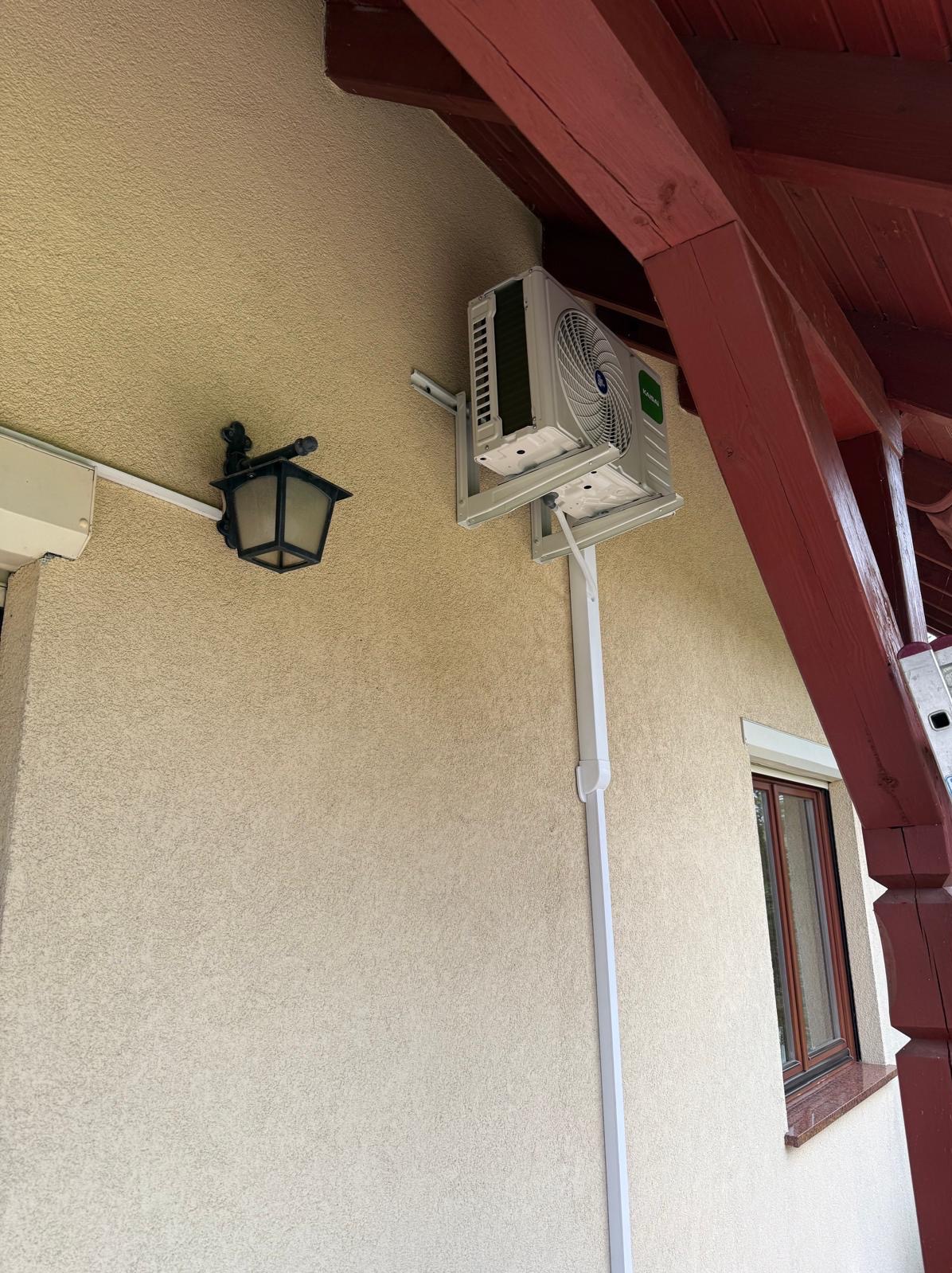 Zewnętrzna jednostka klimatyzacji zamontowana na elewacji budynku pod drewnianym zadaszeniem. Widoczna lampa zewnętrzna i rura odprowadzająca skropliny.