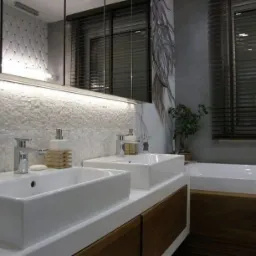 Nowoczesna łazienka z dwoma białymi umywalkami nablatowymi, chromowanymi bateriami, mozaiką na ścianie, podświetlanym lustrem, drewnianymi żaluzjami i dekoracyjnym rysunkiem na ścianie.